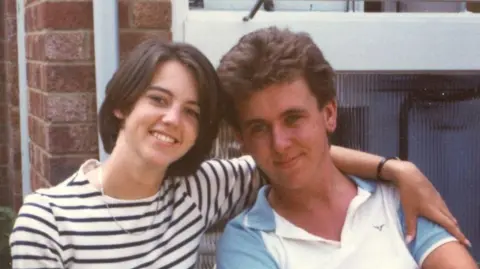 Karen Goddard An old photo of Karen Goddard with her arm around her husband Tim. She has a brown bob and wears a white long sleeved top with black stripes and a necklace around her neck. Tim has brown hair and wears a white polo top with blue short sleeves.