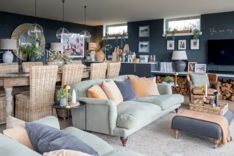 IWC Media A stylish open-plan living and dining area features soft green sofas, woven chairs, and layered neutral decor against dark painted walls.
Glass pendant lights hang above a wooden dining table, while shelves, plants, and framed photos add warmth and personality.
