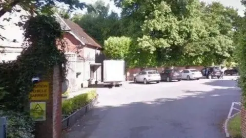 A Google street view image of the trust facility in Tadworth. There is a line of cars parked under a large tree beyond a bricked entrance tot he paved area