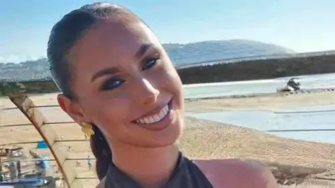 Erin has brown hair tied back and smiles at the camera. There is a beach behind her