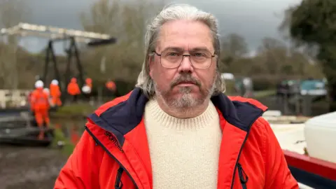 A man with long grey hair slicked back, grey facial hair and glasses. He is wearing a cream jumper and a red waterproof coat