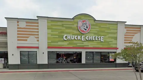 Google The exterior of a large building. There is a large green sign which has a cartoon like picture of a mouse and reads 'Chuck E Cheese'.