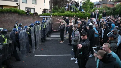 A line of riot police face off against a large group of protestors in the middle of a road. 