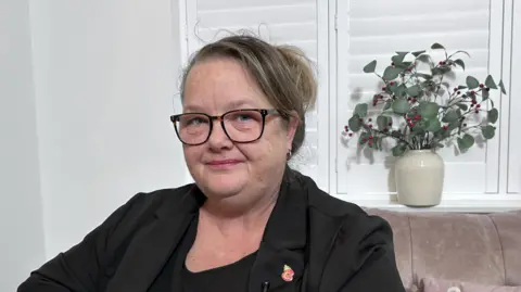BBC/Kate Bradbrook Woman with dark blonde hair and glasses sits on a sofa. She is wearing a black top and blazer.