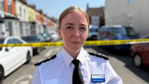 Ch Insp Nicola Davis-Lyons, who is standing in the middle of a street with terraced houses and cars on either side. Her fair hair is pulled back from her face and she is wearing a white police uniform shirt with epaulettes on her shoulders and a black tie. Behind her is a line of yellow police tape.