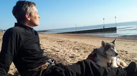 An image of Wayne Dixon and his dog Koda sitting on a sandy beach with the sea in the background. Koda is a Northern Inuit breed and is laying down with his eyes closed next to Wayne. Wayne is dressed in all black and is looking out to sea