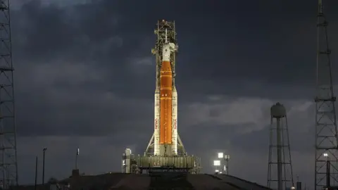 Getty Images Orange rocket with white tip stands vertically ready to lift off lit up by flood lights.
