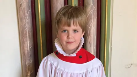 George smiles at the camera wearing white and red chorister robes. He has blond hair, some of which hangs over his forehead.