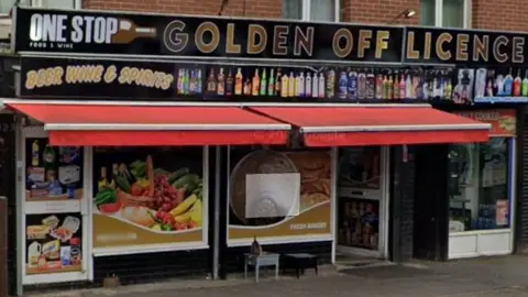 Large shop front with red awning. 
Pictured above the awning are bottles of alcoholic and non alcoholic drink. 
and the shop name : One Stop: Golden Off Licence, beer wine and spirits. 
Windows and doors are covered in images of general groceries (milk, eggs, fruit and vegetables).