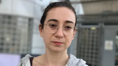 A woman wearing a grey hoodie and glasses looks at the camera.