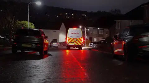 The rear of a police van, with red lights glowing off a wet road surface middle of shot, flanked by cars and houses on each side.