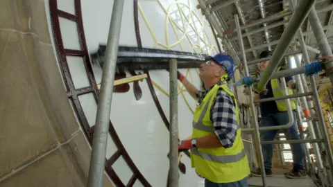 BBC Repair Shop host Steve Fletcher peers at the clock face at Manchester Town Hall, while wearing a green high-vis vest and a blue hardhat. 
He is stood on a scaffold with another worker. 