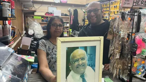 A woman and a man standing behind a photo of a man smiling in a suit with a shop behind them
