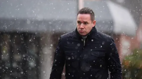 A man with short dark hair and wearing a dark jacket walks through falling snow.