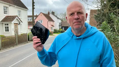 Henry Godfrey-Evans/BBC Paul Maleary wears a light blue hooded jumper, and stands at the side of a road with houses behind. He holds up a wing mirror from a vehicle.