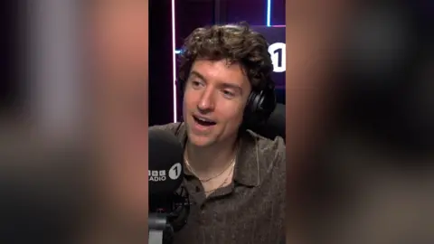 A man in a brown shirt speaks into a radio 1 microphone.