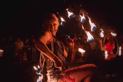 Kitt Bunney A women is holding a leaf shaped piece of metal that is on fire. She is side on facing the camera with her front knee lifted up. 