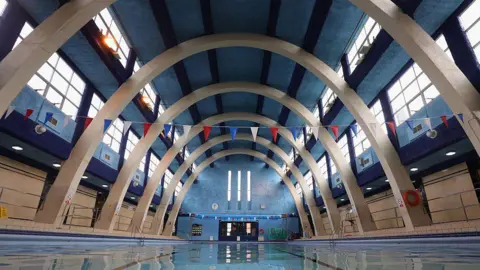 A swimming pool with parabolic arches. the ceiling is painted blue with four dark blue lines down it. There is bunting running across the pool and windows either side