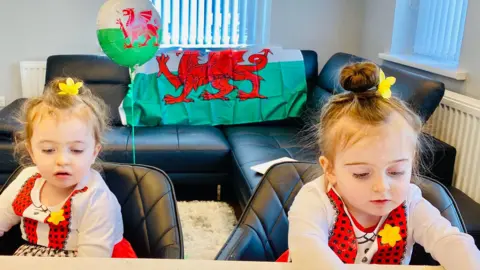 Family photo Twin eight-year-old girls wearing Welsh lady costumes of white and red tops and red skirts, with daffodils in their tied up hair, sit at a kitchen island looking down. A Wales flag is on display on a sofa in the background, and next to it is a helium balloon which also has a Welsh flag design on it.