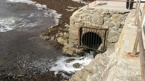 BBC A pipe at a beach in Cornwall. The pipe is coming from beneath a platform overlooking the sea. The hole is covered by a metal grate and liquid is pouring out of the pipe and into the sea.