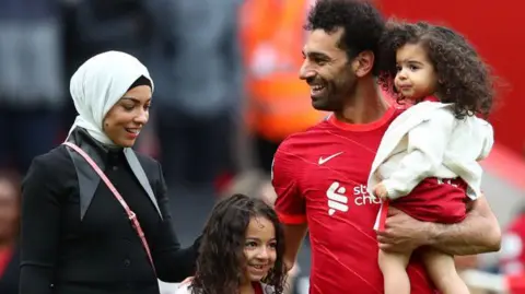 Getty Images Mohamed Salah holds his youngest daughter Kayan in his arms. His wife Magi and their eldest daughter Makka are standing next to him during end-of-the-season celebrations in 2022 at Anfield.