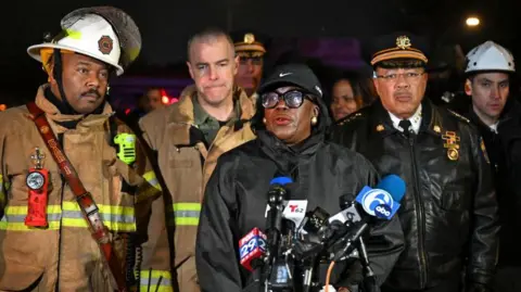 Getty Images Philadelphia Mayor Cherelle Parker wears a black Nike baseball cap and a black raincoat as she speaks to reporters at the site of the crash, flanked by fire crews and police in uniform