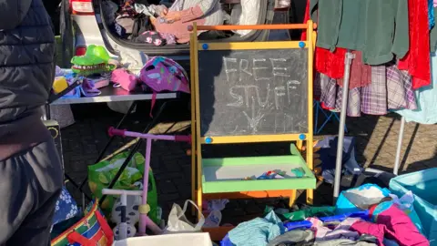 A large amount of clothes and children's toys - including a blackboard reading "free stuff" is sprawled out across the floor, on a foldable table, and in a car boot at a car boot sale in the winter sunshine.