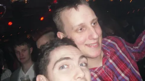 An old selfie-style image of Lee Walker and his friend in a nightclub. He is wearing a red and purple check shirt and is looking away from the camera. His friend is looking directly into the camera. Another man can be seen in the background looking startled while looking into the camera. 