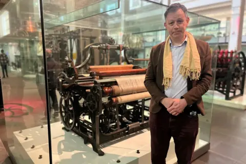 Ben Russell wearing a brown suit over a off-white shirt with a yellow scarf around his neck stands next to a cotton loom inside a large glass cabinet