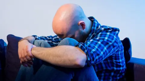 A man is hunched over on a sofa with his head between his knees and his arms locked around them.
