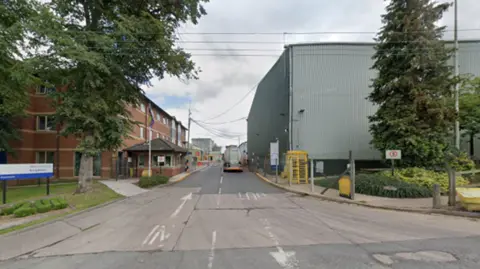 An entrance to a factory with a red brick, three-storey, building on the right and the grey factory on the left. 