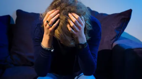PA Media A woman is sat with her head in her hands on a dark blue sofa. She has dark blonde hair and silver rings on her fingers.