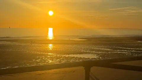 BBC The sun setting on a low tide at St Ouen's bay with the handrail in the foreground and a small boat to the left in the ocean. People walking along the beach.