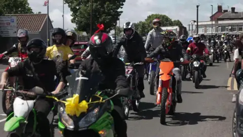 Dozens of motorcyclists travel down a road. The bike at the front has a yellow ribbon over the headlights.