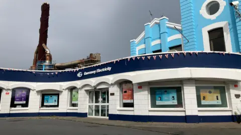 BBC Exterior of the Guernsey Electricity main office. It is a blue and white building with several windows and a set of glass doors. It is a gloomy day and there is a brown industrial chimney in the background.