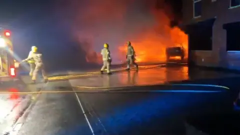 Five firefighters working together to get a hose to a vehicle on fire near a building