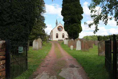 Billy McCrorie A view up a pathway past gravestones and trees to a white church building with a spire.