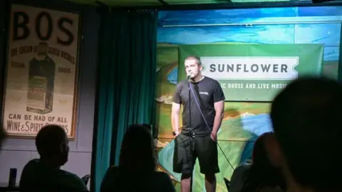 Jonathan Campbell Cathaóir stands on stage in front of a mic. He has buzzed hair, wearing a black t-shirt and shorts. An old fashioned advert poster for Bos whiskey is to his left, and behind him is a sign for the venue called Sunflower, located in Belfast.