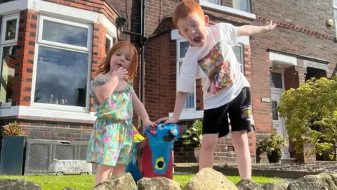 James Bowman A young girl and young boy stand either side of a brightly coloured Superlambanana in their garden.