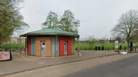Google A hexagon-shaped block of public toilets on the edge of a green park, next to a road. The building has an overhanging green roof, and surrounds of the doors are coloured red, orange and yellow.
