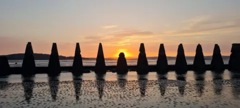 Jim Kerr A row of concrete pylons on a beach, with the sun setting behind the smaller middle pylon