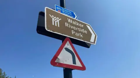 A brown and white sign for Walker Riverside park above a red triangle sign warning of a sharp bend.
