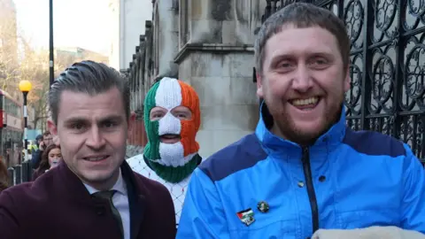PA Media Lawyer Darragh Mackin wearing a maroon coat arrives with DJ Provai, in a tricolour mask and white jumper and Dan Lambert in a blue coat. 