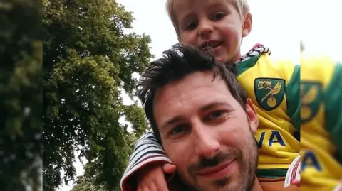 Finnbar's Force A man with black hair and a beard smiling with his young son resting on his shoulders, wearing a Norwich City shirt.