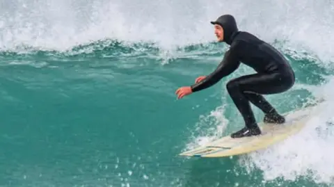 @rusty.uk A surfer in a hooded wetsuit takes a right hand wave on a surfboard