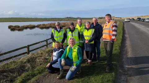 Froglife A group of eight people, two kneeling, are gathered by a wooden fence with a pond to the left and a road to the right. They are wearing hi-vis vests. In the distance Bamburgh Castle can be seen on the horizon.