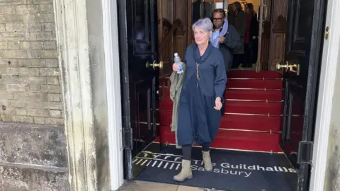 Caroline Sturgess leaving the Guildhall in Salisbury. She is wearing a navy dress and cardigan and is holding a water bottle. She has short purple hair.