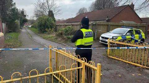 White and blue police tape is stretched across a yellow metal gate near an alleyway between houses. You can see the back of a police officer with a high vis vest and black helmet and a police car is parked to the right.