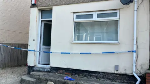 A police cordon outside a white building. On a step is a box of blue gloves.