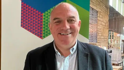 A smiling man looking straight ahead. He is wearing a grey suit jacket with a light blue shirt underneath.